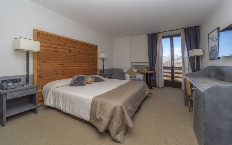 Camera d'hotel serena con letto in legno, mobili grigi e vista su un balcone con alberi e cielo blu.