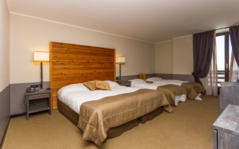 Hotel room with two beds, beige walls, tan carpet, nightstands, lamps, and large windows with dark curtains.