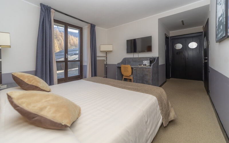 Serene hotel room with a double bed, a second bed, gray curtains, mountain view, desk with yellow chair, and a TV.