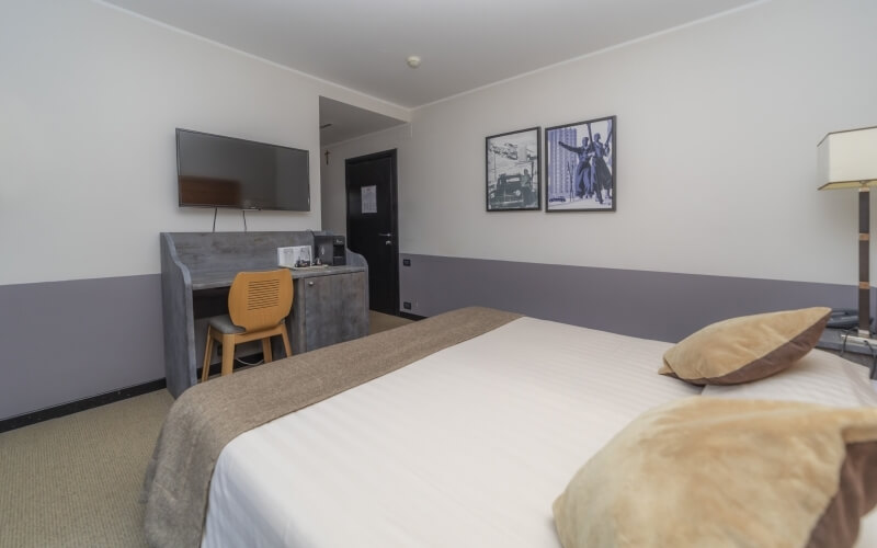 Camera d'hotel con letto, scrivania, sedia gialla e TV a parete, decorata in bianco e grigio con moquette beige.