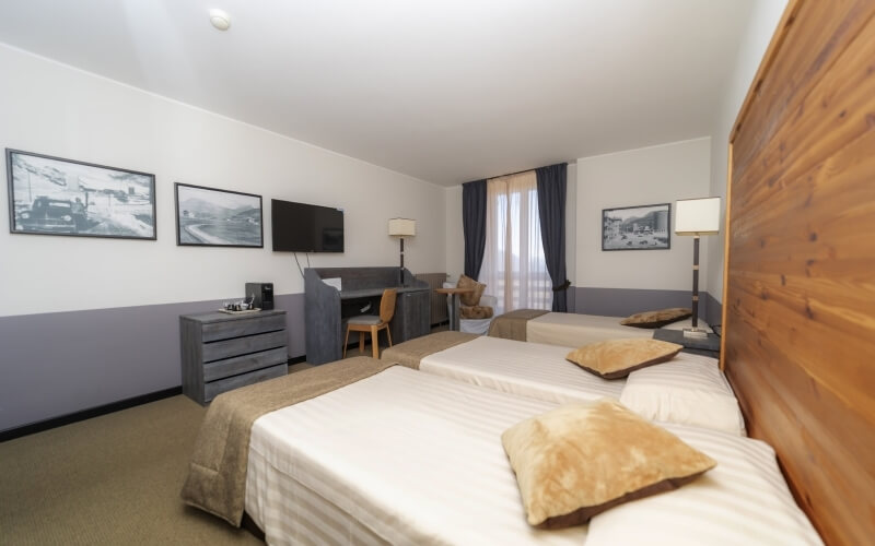 Hotel room with two beds, wooden dresser, flat-screen TV, desk by window, and black-and-white photographs on the wall.