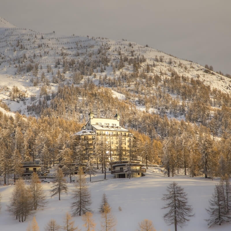 Serene winter scene with a snow-covered mountain, a large yellow hotel, and trees in a peaceful, overcast atmosphere.