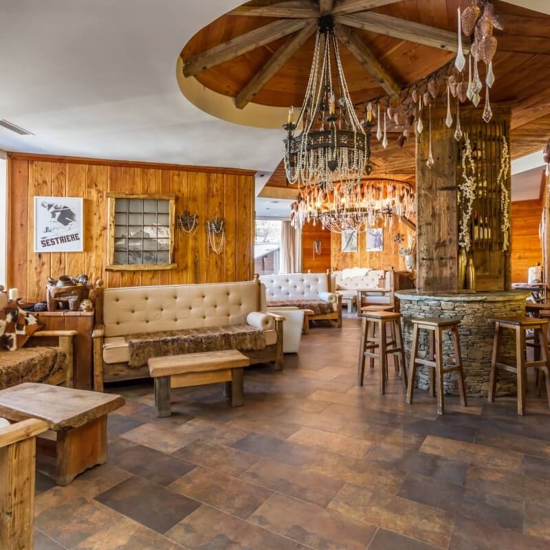 Rustic room with a stone bar, wooden stools, cozy seating area, exposed beams, chandelier, and natural light.