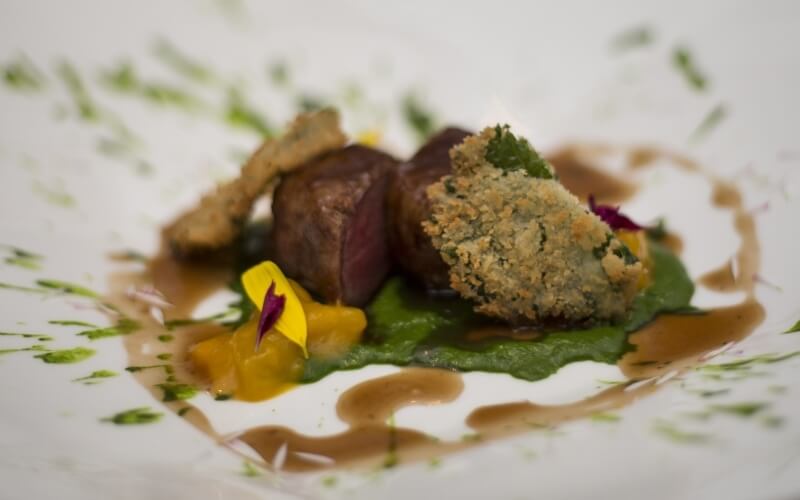 Close-up of hand-torn beef with sauces, colorful vegetables, and artistic elements on a vibrant plate against a white background.