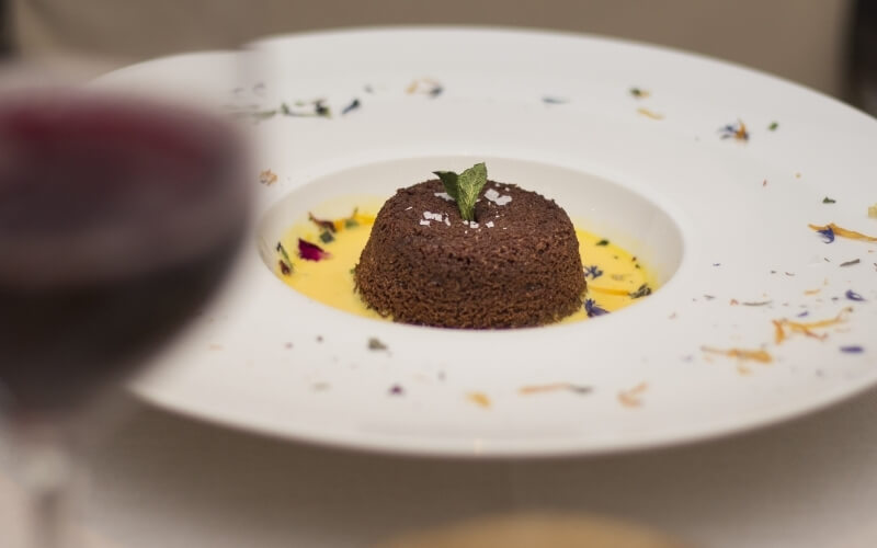 Dessert on a white plate with a brown cake in the center, surrounded by colorful flower petals in a blurred table setting.