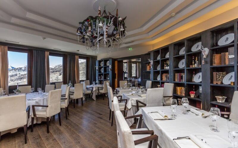 Formal dining room with tables set in white linens, dark wood furniture, chandelier, bookshelves, and windows.