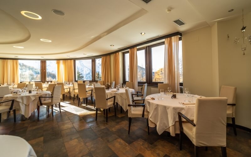 Elegant dining room with round tables, white tablecloths, and a wall of windows, featuring a curved ceiling and warm tile floor.