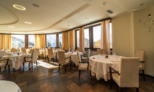 Elegant dining room with circular tables, white linens, beige chairs, and natural light from narrow windows with orange curtains.