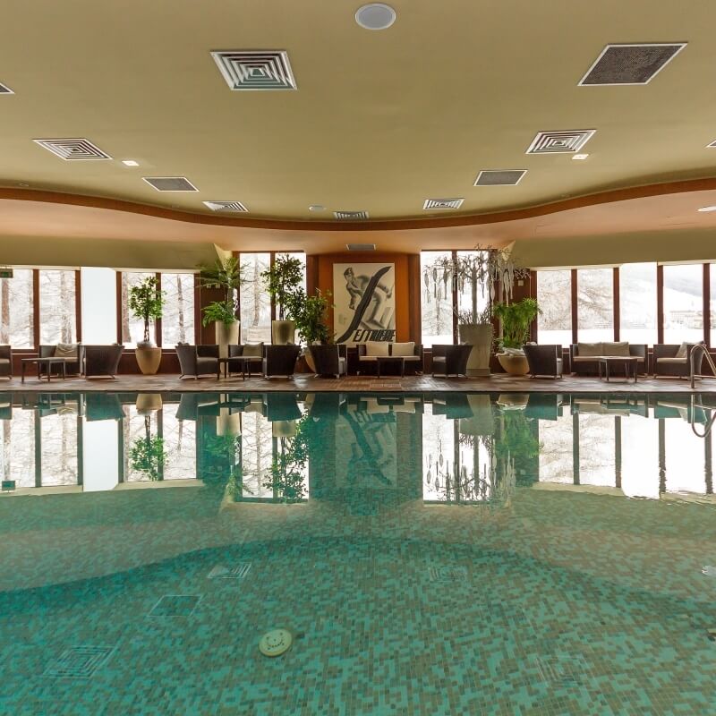Indoor pool with reflective dark greenish-blue surface, surrounded by lounge chairs, potted plants, and light brown walls.