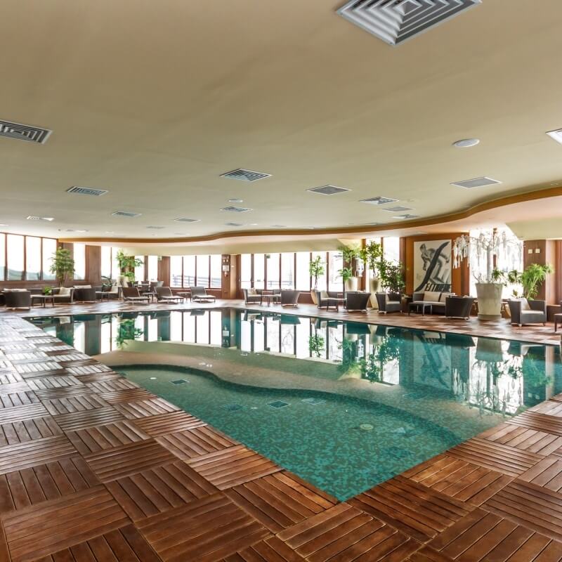 Indoor swimming pool with lounge chairs, tables, and a hot tub, surrounded by windows and potted plants in a luxurious setting.