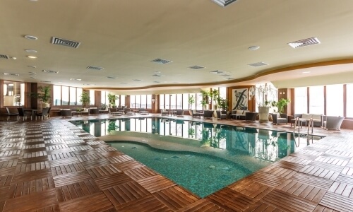 Indoor pool with blue-green water, surrounded by lounge seating, plants, and large windows in a spacious, bright room.