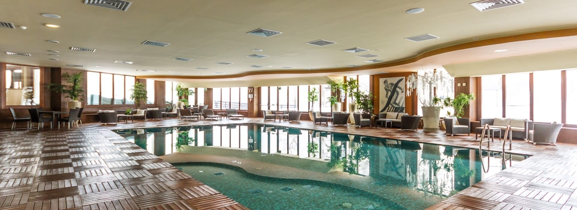 Indoor swimming pool with dark green tiles, wooden deck, contemporary seating, and large windows letting in natural light.