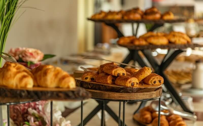 A table displays an assortment of pastries: croissants, chocolate-filled croissants, and tiered pastries in a cafe setting.