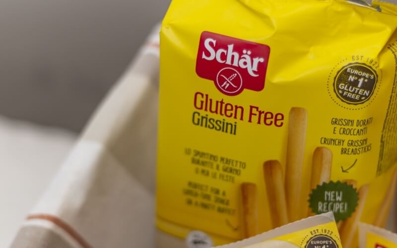 Yellow triangular bag of Schar gluten-free grissini with red logo, "Gluten Free" text, and circular black certification logo.