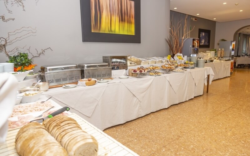 Buffet table with sliced meat, plates, decorative plants, and a large muted painting in a banquet hall setting.