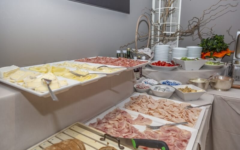 Buffet table with sliced meats, condiments, and fresh toppings, set for a catering event, against a light gray wall.