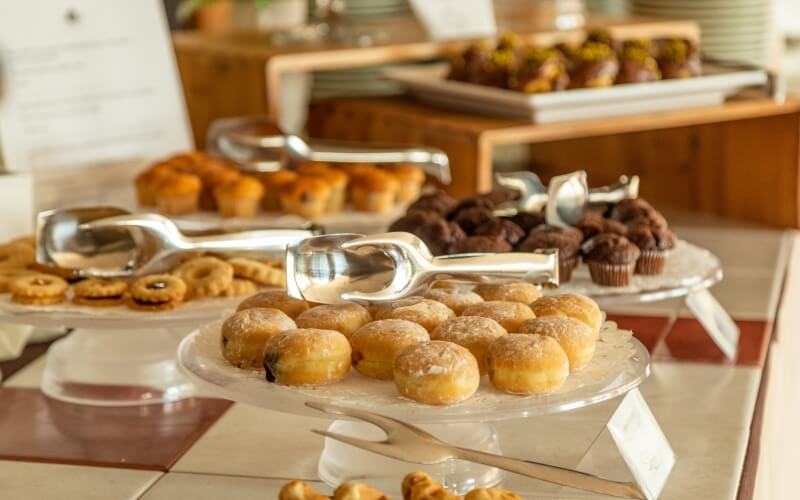 Buffet table with assorted desserts: donuts, muffins, and cookies on glass dishes, with silver utensils and name cards.