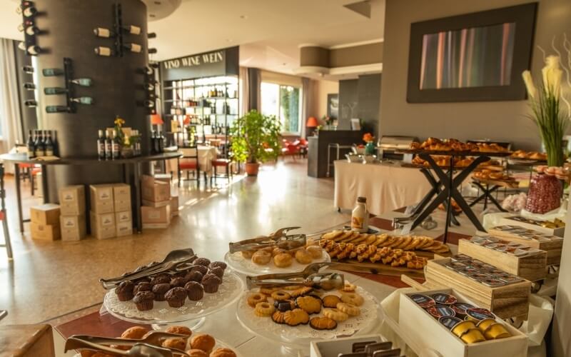 Buffet table with pastries, chocolates, and cookies in a well-lit room with a bar area and large windows.