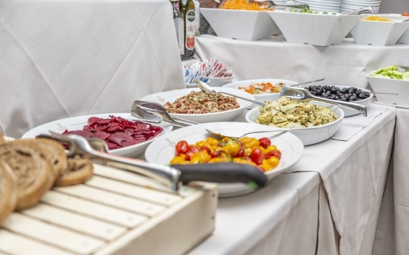 Buffet table with assorted dishes, including meats, salads, and fruits, set on a white tablecloth with an empty chair.