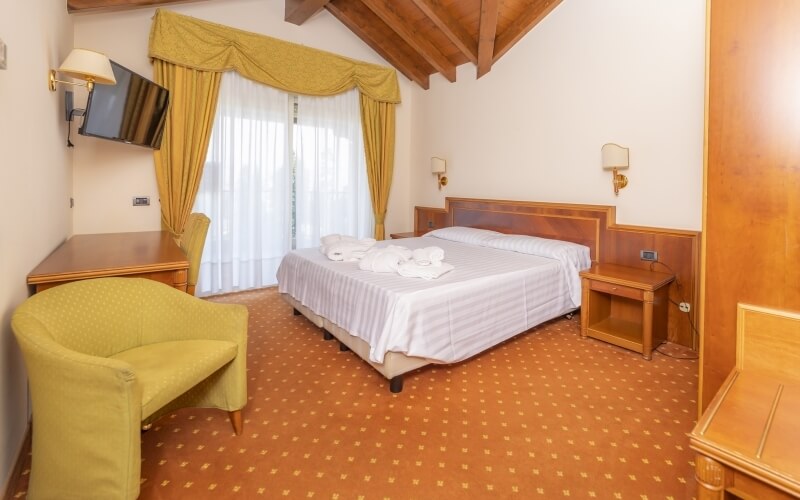 Cozy hotel room featuring a double bed, wooden furniture, a mounted TV, yellow curtains, and a sloping ceiling with beams.