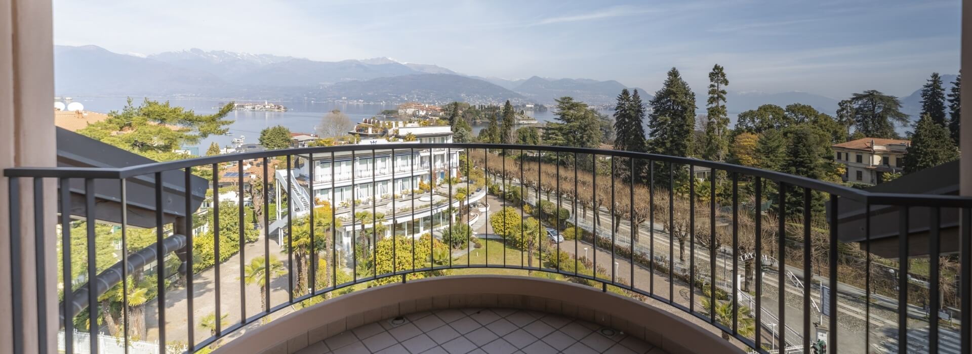 Balcony with black railing overlooking a serene landscape, water, trees, buildings, and distant mountains under a clear blue sky.