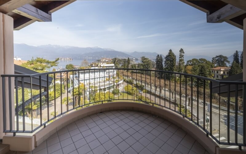 Serene balcony view with tiled floor, black railing, trees, buildings, water, mountains, and a blue sky with clouds.