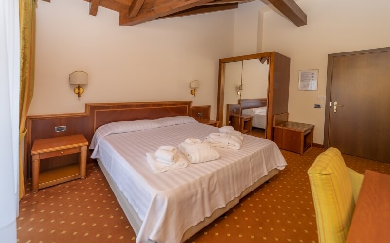 Cozy hotel room featuring a bed with white linens, nightstands, a mirror, a bench, and a yellow chair on patterned carpet.