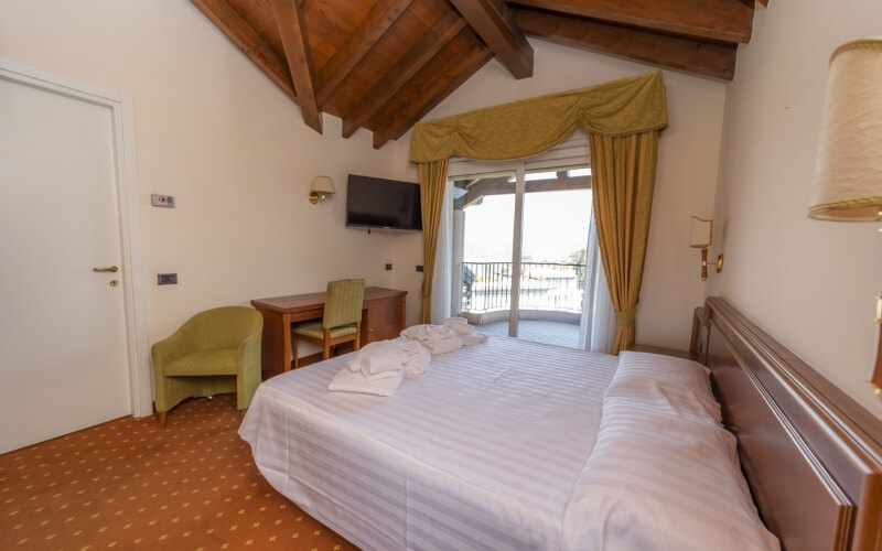 Cozy hotel room with a large bed, desk, chair, TV, balcony window, and wooden beam ceiling, featuring red carpet flooring.