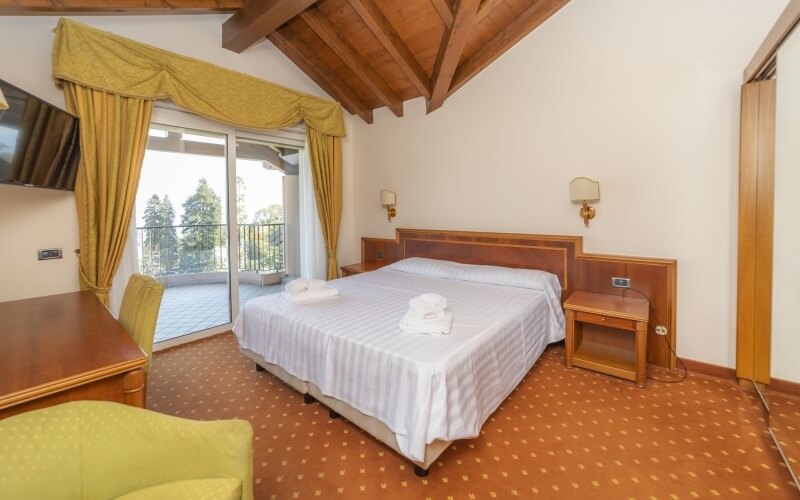 Cozy hotel room with a double bed, balcony, desk, and rustic decor featuring a patterned carpet and exposed beam ceiling.