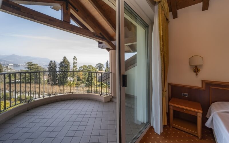 A room split view: cozy interior with a bed and balcony overlooking trees and mountains under a clear blue sky.