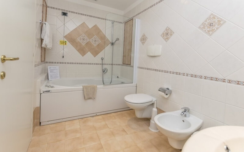 A clean bathroom featuring a white bathtub, toilet, bidet, sink, beige tiled floor, and decorative white tiled walls.