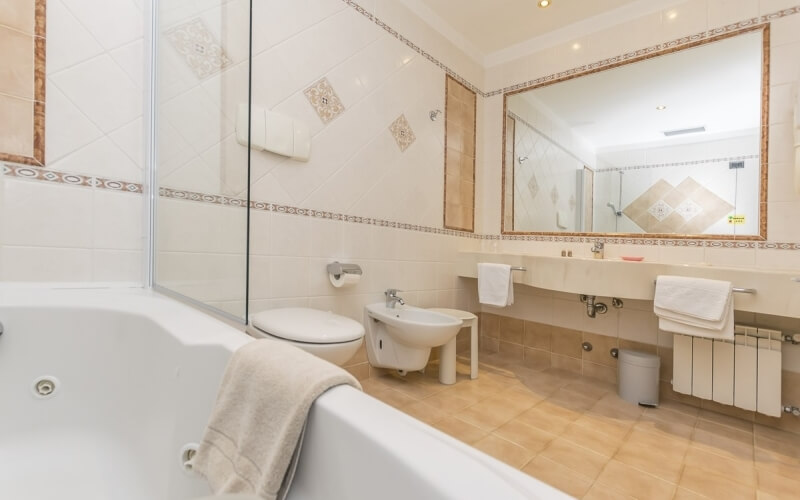 Modern bathroom with white and tan elements, featuring a bathtub, bidet, toilet, sink, towel rack, and tile flooring.