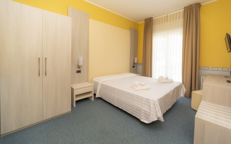 Hotel room with yellow walls, blue carpet, white bed with towels, nightstand, closet, and bathroom area with sink and toilet.