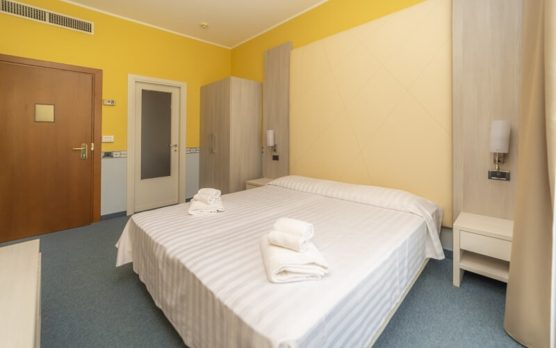 Cozy hotel room with a double bed, nightstand, closet, and cheerful yellow walls, featuring a light blue carpet.