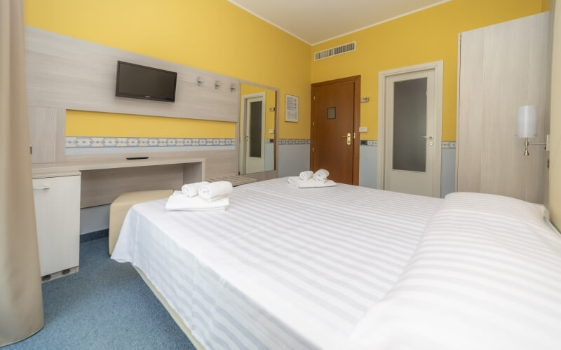 Bright hotel room with a large bed, TV, wooden desk, and bathroom door, featuring yellow walls and blue carpeting.