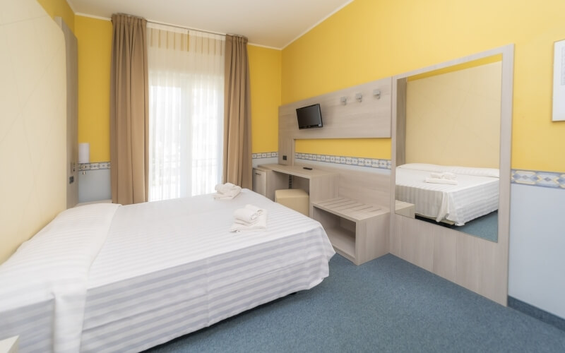 Small hotel room with yellow walls, blue carpet, large bed, desk with TV and mini-fridge, and a mirror reflecting the space.