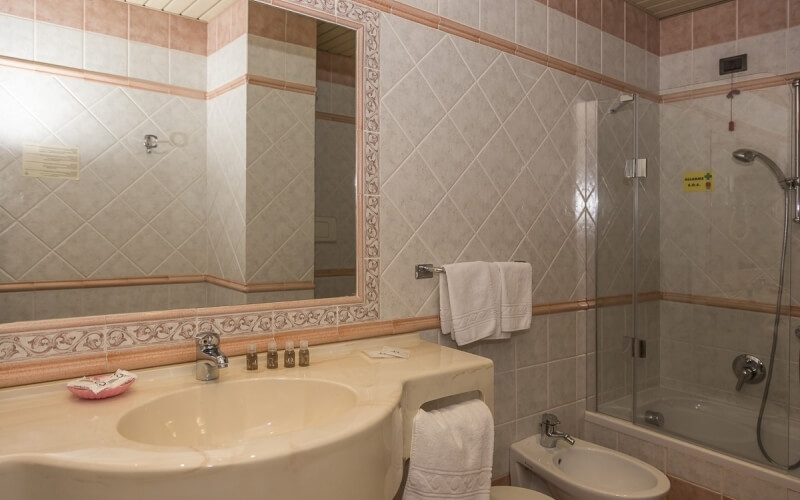 A clean bathroom featuring a beige and white color scheme, large mirror with brown floral border, sink, and glass-enclosed shower.