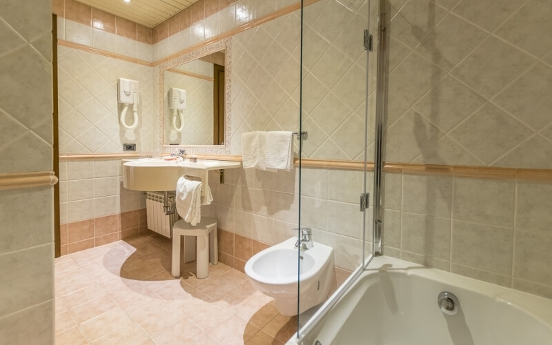 A clean bathroom with beige and brown tiles, featuring a sink, toilet, bathtub, and towels on a stool.