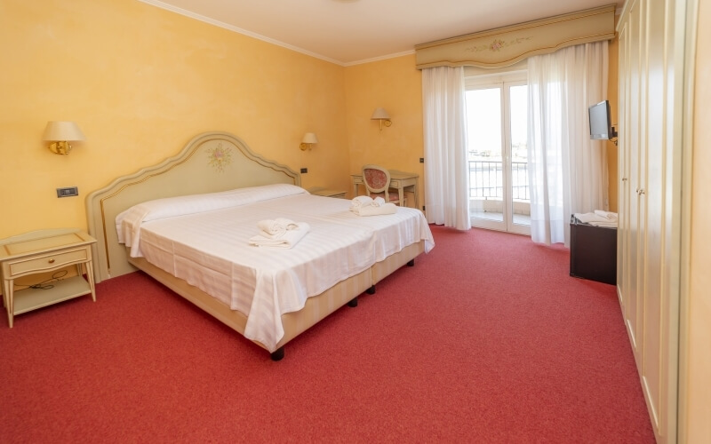 Cozy hotel room with a bed, nightstands, desk, warm lighting, and a large window with sheer curtains.