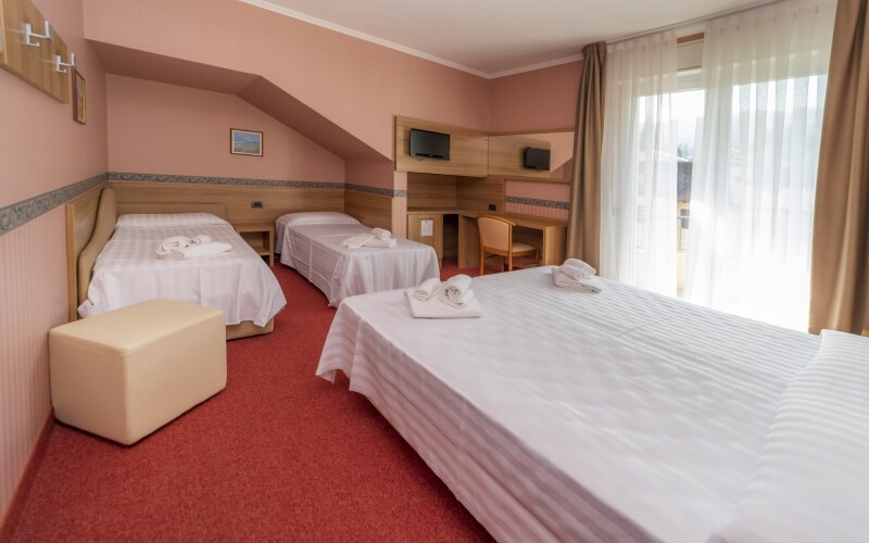 Hotel room with three beds, light pink walls, red carpet, desk, and natural light from a window, exuding comfort and cleanliness.