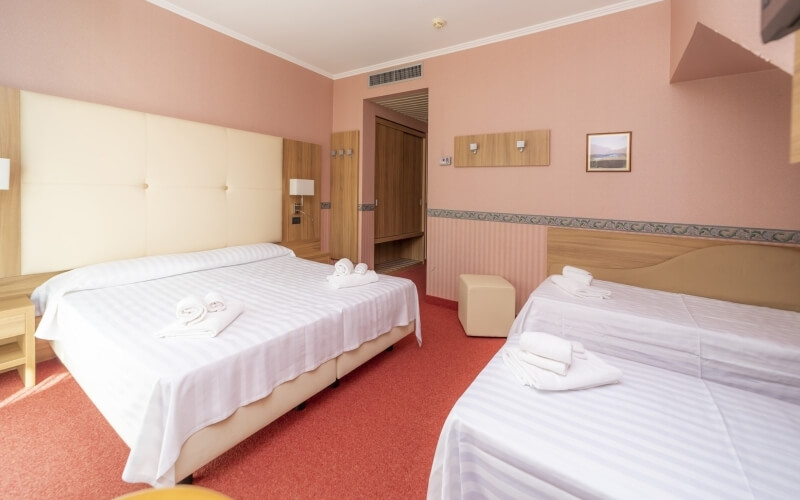 Serene hotel room with two beds, peach and wooden headboards, beige ottoman, nightstands, and light pink walls.