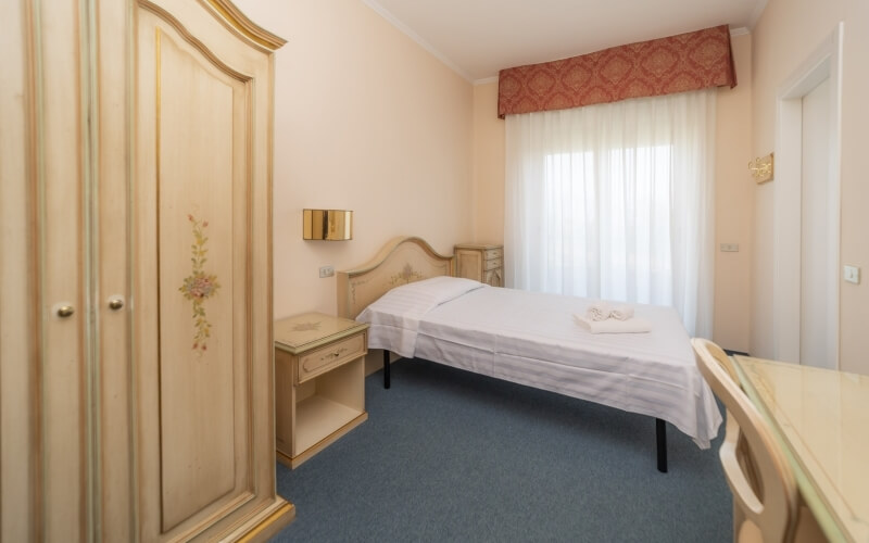 Cozy cream-colored bedroom with a single bed, floral nightstands, a wardrobe, and a desk, illuminated by natural light.