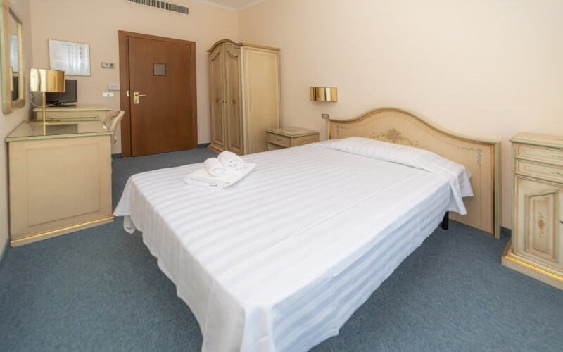 Hotel room featuring a bed with white sheets, nightstands, a wardrobe, blue carpet, and beige walls.