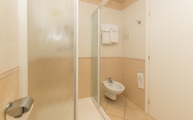 Small bathroom with peach tiles, shower, sink, toilet, and white towels on a rack; tan tiled floor.