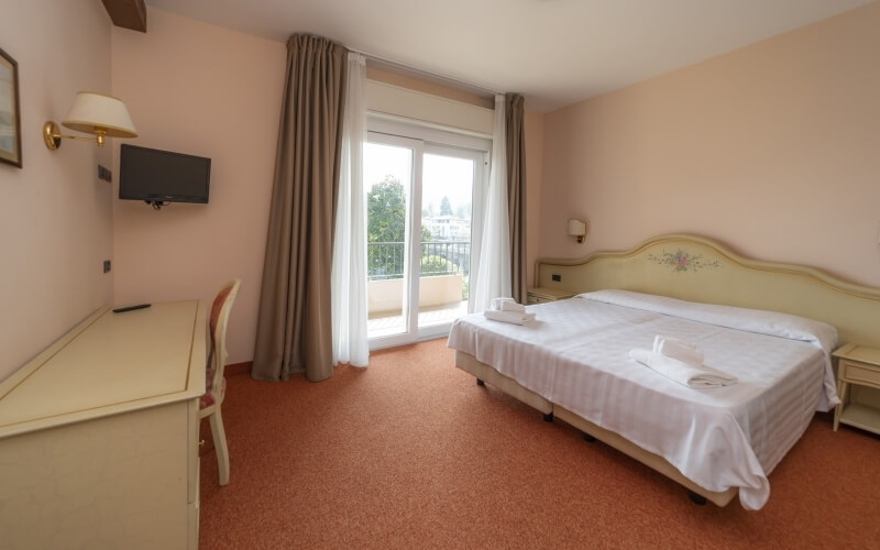 Hotel room with light walls, a bed with white comforter, desk, lamp, TV, and balcony window, creating a serene atmosphere.