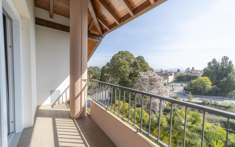 A serene second-floor balcony with a metal railing, wooden roof, and tiled floor, surrounded by greenery under a clear blue sky.