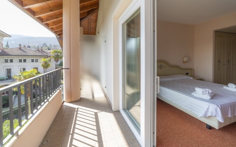 Juxtaposition of a balcony with palm trees and mountains outside, and a cozy bedroom with a bed and orange carpet inside.