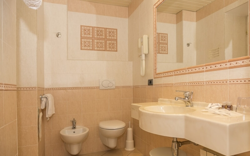 A clean bathroom featuring a sink, toilet, bidet, decorative tiles, mirror, and towels, with toiletries on the counter.