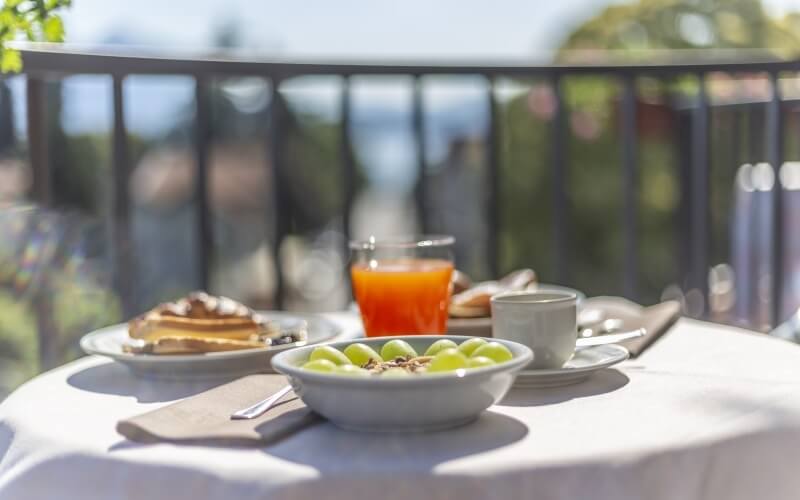 Outdoor breakfast table with grapes, coffee, orange juice, and waffles, set against trees and water in a serene setting.