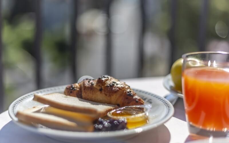 Close-up of a plate with buttered bread, croissant, honey, and dark sauce, with a glass of orange juice outdoors.